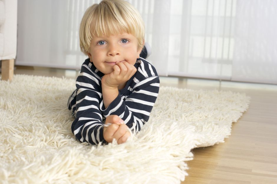 A young child with blonde hair and blue eyes, wearing a navy and white striped long-sleeve shirt, is lying on their stomach on a plush, cream-colored shaggy carpet in a well-lit living room with light