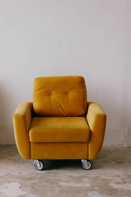 Close-up of a person using a handheld vacuum cleaner to perform surface cleaning on an olive green, fabric-upholstered armchair, part of a living room setting that includes a dark gray sofa in the bac